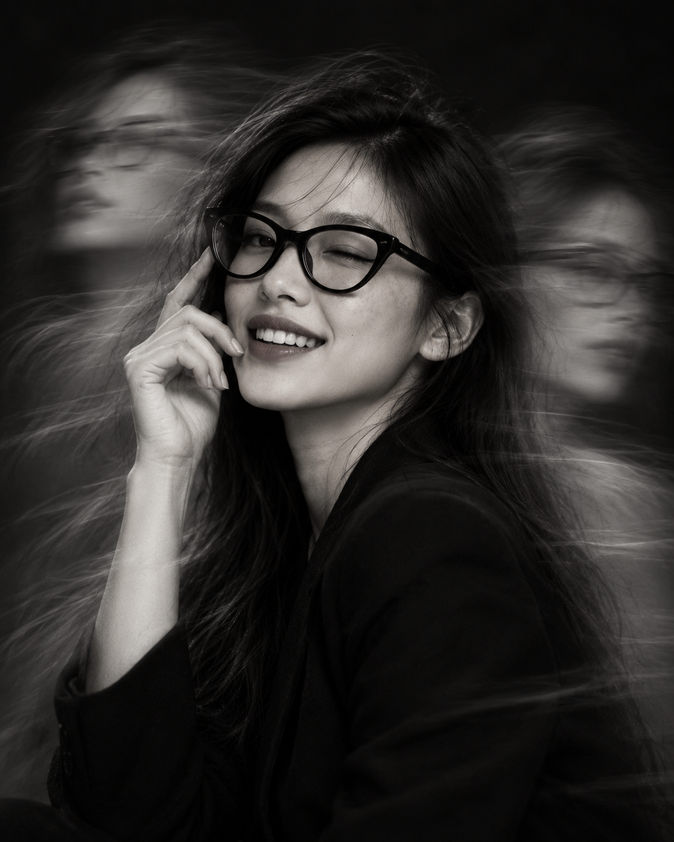 Mulher de óculos sorrindo com a mão no rosto, em fotografia preto e branco com efeito de movimento